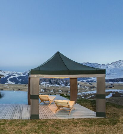 Grün-graues Lodenzelt steht neben dem Pool in der Abenddämmerung. Darunter stehen 2 Holzliegen.
