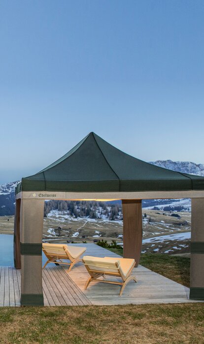 A Loden tent in the colours grey and green is located beside the pool at sunset. Under it are 2 wooden benches. 