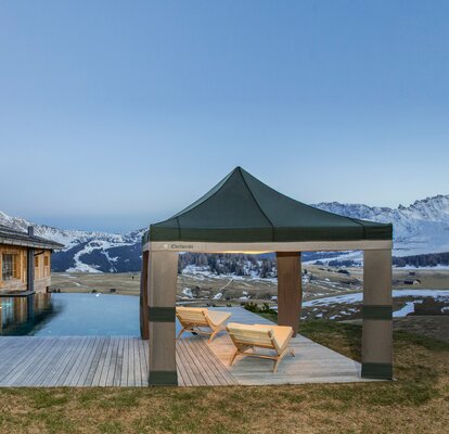 A Loden tent in the colours grey and green is located beside the pool at sunset. Under it are 2 wooden benches. 