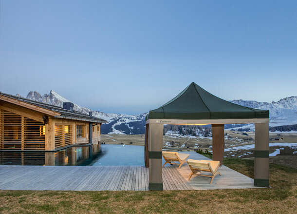 A Loden tent in the colours grey and green is located beside the pool at sunset. Under it are 2 wooden benches. 