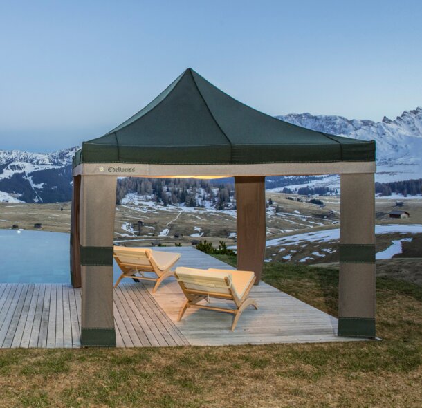 Grün-graues Lodenzelt steht neben dem Pool in der Abenddämmerung. Darunter stehen 2 Holzliegen.