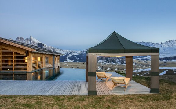 A Loden tent in the colours grey and green is located beside the pool at sunset. Under it are 2 wooden benches. 