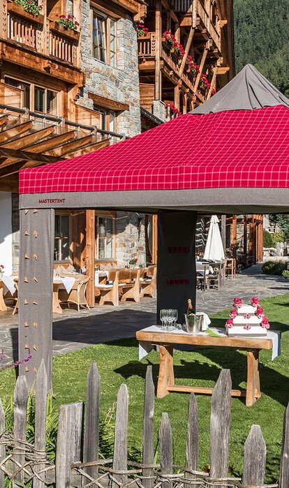 A loden tent in the colours red and grey is located in the garden of a restaurant; under the gazebo is a table with champagne glasses and cake. 