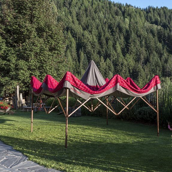 Halbaufgezogener Faltpavillon mit rot-grauem Lodenstoff-Überzug im Garten.