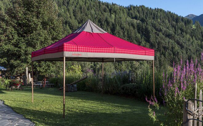 The gazebo with the red-grey loden fabric cover has been fully opened. It is placed in the garden. 