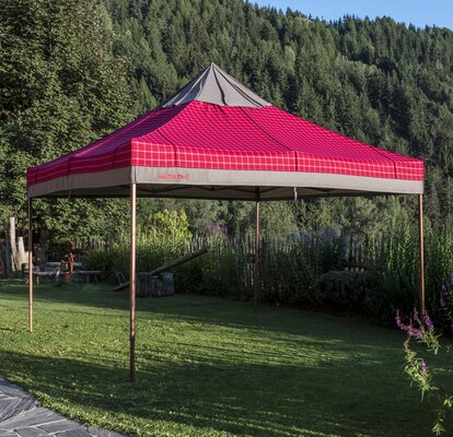 The gazebo with the red-grey loden fabric cover has been fully opened. It is placed in the garden. 