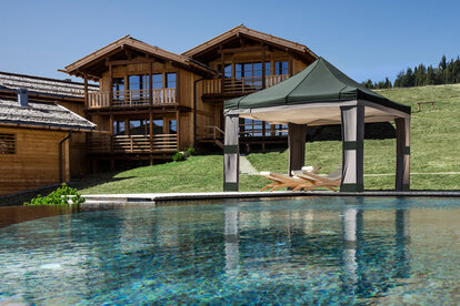 Neben dem Schwimmbad ist ein Lodenzelt mit Himmel. Unter dem Lodenzelt sind zwei Liegestühle.