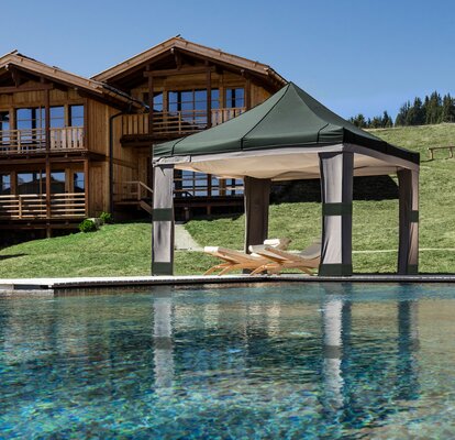 Neben dem Schwimmbad ist ein Lodenzelt mit Himmel. Unter dem Lodenzelt sind zwei Liegestühle.