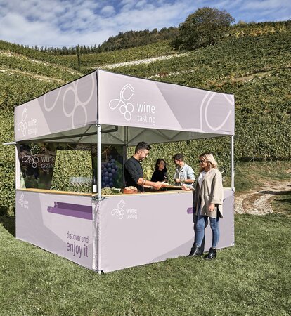 Un gazebo pieghevole con tetto piano in mezzo alle vigne. Un somelier sta servendo vino e formaggio a degli ospiti.