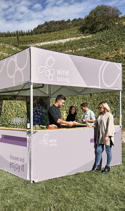 Un gazebo pieghevole con tetto piano in mezzo alle vigne. Un somelier sta servendo vino e formaggio a degli ospiti.