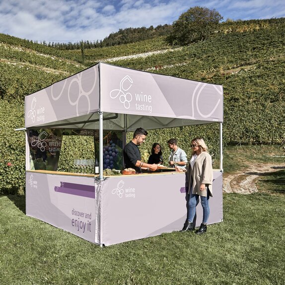 Un gazebo pieghevole con tetto piano in mezzo alle vigne. Un somelier sta servendo vino e formaggio a degli ospiti.