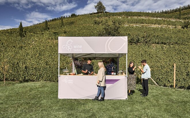 Simple printed gazebo with flat roof and the lettering "wine tasting". The owner of the vine yard is presenting the wine to a client, while two other people are already tasting the wine. In the background: vineyard with vines. 