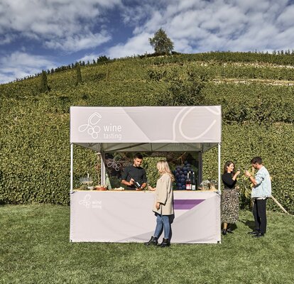 Simple printed gazebo with flat roof and the lettering "wine tasting". The owner of the vine yard is presenting the wine to a client, while two other people are already tasting the wine. In the background: vineyard with vines. 