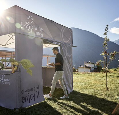 Simple printed gazebo with flat roof and the lettering "wine tasting" is located on a meadow with apple trees. The owner of the wine yard is entering the gazebo through the rolling door of a sidewall.