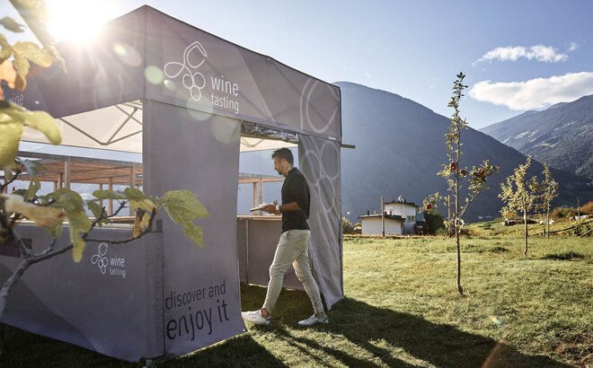 Simple printed gazebo with flat roof and the lettering "wine tasting" is located on a meadow with apple trees. The owner of the wine yard is entering the gazebo through the rolling door of a sidewall.