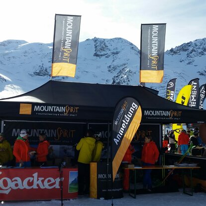 Schwarzes Zelt bei einem Wintersport-Event im Schnee. Viele Besucher in bunter Skibekleidung stehen rund um das Zelt, im Hintergrund schneebedeckte Berge und diverse Markenfahnen.