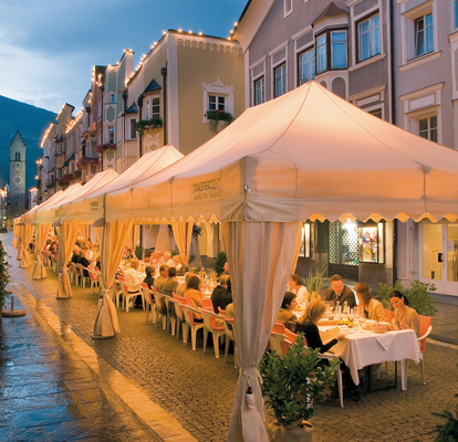 Set di gazebo esterni eleganti 4x8 m ecru per le vie di Vipiteno per una cena - Mastertent Set di gazebo esterni ecru eleganti 4x8m kit Royal con tende drappeggiate e frontalino ondulato per cena di lusso nella città di Vipiteno di sera con le luci Mastertent