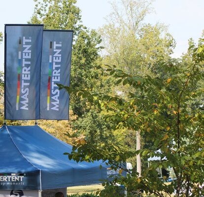Blue peak flags in a row on top of canopy tents printed with the Mastertent logo set in a wooded area.