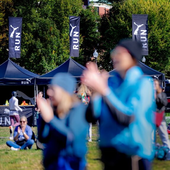 Blurred figures clapping in the foreground in front of three, black 10x10 Mastertents equipped with peak flags and printed with a white Puma logo.