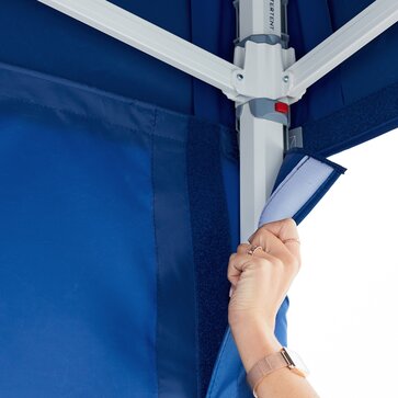 A woman's hand is currently fastening the blue sidewalls with velcro strips along the gazebo legs. 